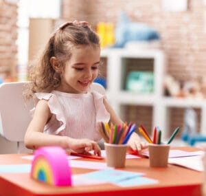 Young girl sitting at classroom table drawing with colored pencils for kindergarten readiness assessment