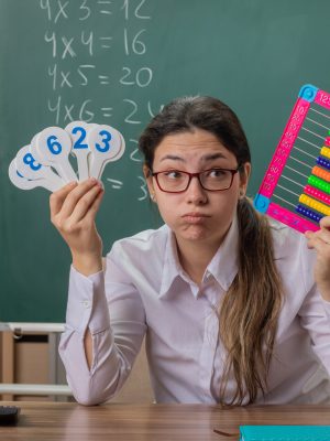 young woman teacher wearing glasses sitting at school desk with bills and number plates looking tired and bored blowing cheeks explaining lesson in front of blackboard in classroom